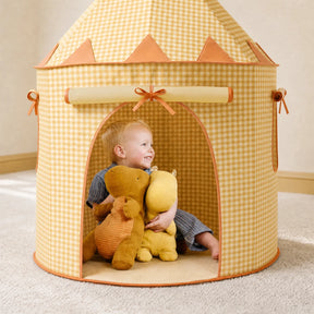 A smiling toddler hugs two plush toys inside the Tiny Land® Kids Tent – Indoor Tent for Toddlers by Tiny Land INC, featuring a yellow checkered pattern, pointed roof, wide entrance, and decorative bows and trim.