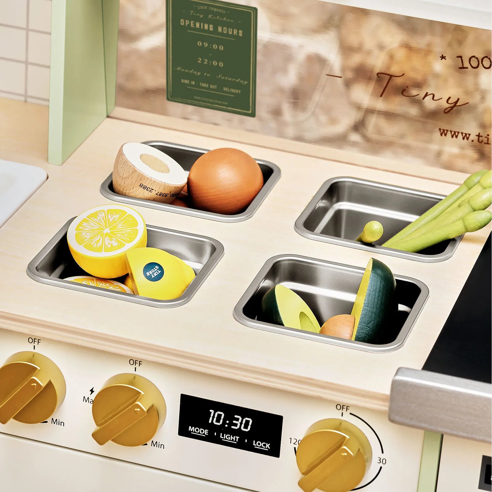 The Tiny Land® BistroSwitch Play Kitchen by Tiny Land features four metal trays with wooden lemon, onion, cucumber, and asparagus. Ideal for pretend play, it has gold knobs and a digital timer display set to 10:30.|Green