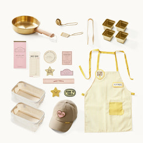 A flat lay of Tiny Land® BistroSwitch Play Kitchen accessories by Tiny Land, including gold tools, mini loaf pans, a yellow-striped apron, beige cap, cloth baskets, recipe cards, paper tags, and star pins on a white background.|White