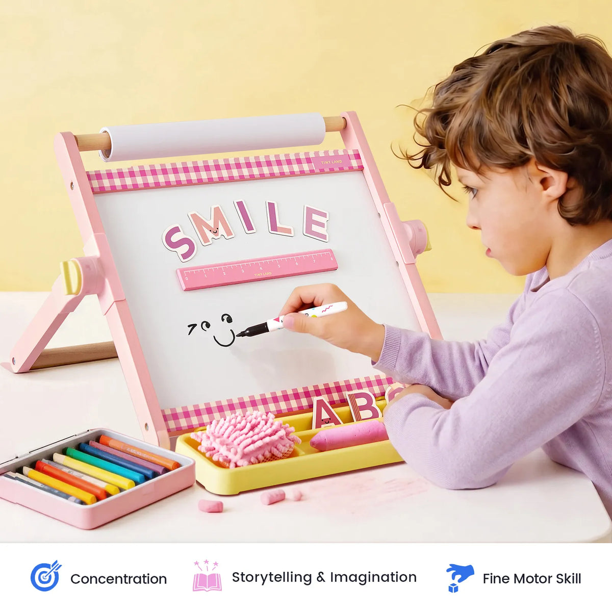A young child draws a smiley face on the pink and white Tiny Land® Double-Sided Magnetic Tabletop Easel, with “SMILE” spelled above. This kids art station by Tiny Land features a magnetic whiteboard, with crayons, chalk, and an eraser nearby. Icons below highlight concentration, storytelling, and fine motor skills.|Pink