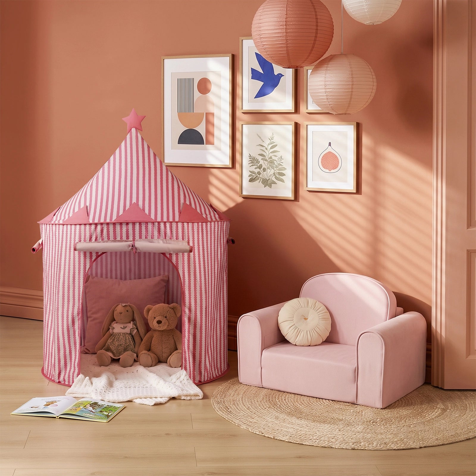 A children’s playroom featuring the Tiny Land® Kids Tent – Indoor Tent for Toddlers by Tiny Land in a pink striped design, paired with two stuffed animals, a pink armchair, a round rug, paper lanterns, and framed wall art set against a peach-colored wall. Sunlight casts soft shadows across the space.|PInk