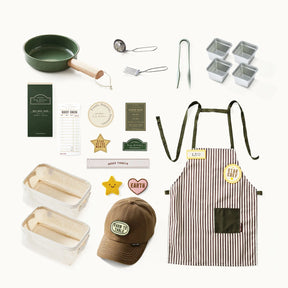 A flat lay of the Tiny Land® BistroSwitch Play Kitchen by Tiny Land features a green pan, whisk, tongs, loaf and square pans, a striped apron, brown hat, order slips, and badges—perfect for pretend play or sparking creativity.|Green