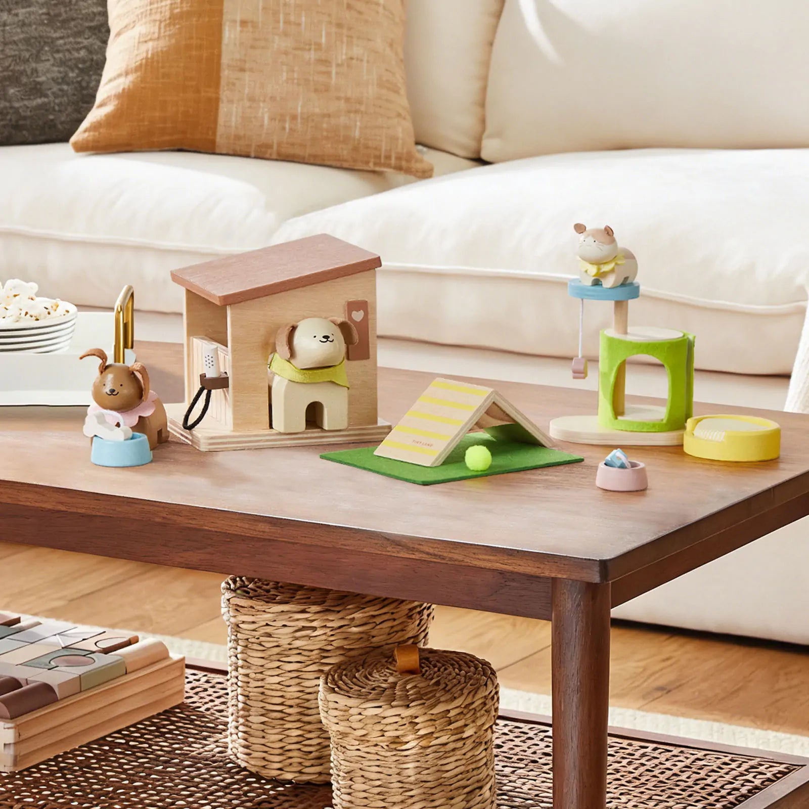 The Early Bird Eggs-Tiny Land® Happy Urban Paws set by Test includes pretend pet toys, a mini house, agility obstacles, food bowls, and animal figurines on a wooden table with woven baskets beneath and a cream sofa in the background.