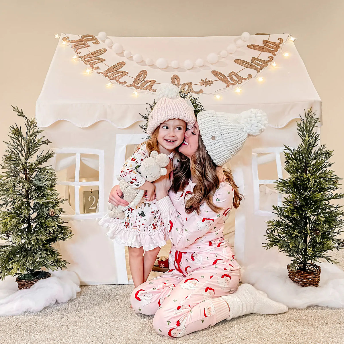 A woman and young girl in festive pajamas smile and embrace by a Tiny Land Bright Play & Art Set—Colorful Easel + Cream Playhouse with Lights—decorated with holiday trees, as the girl hugs a stuffed animal.