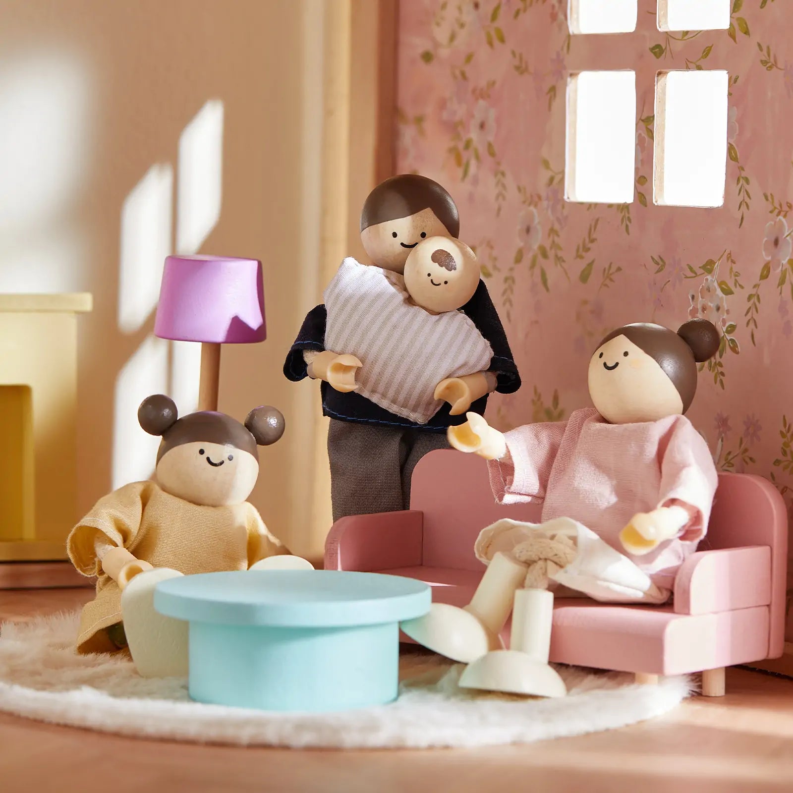 Three Tiny Land® Sweetwood Unicorn Dollhouse dolls with painted faces relax in the cozy living room, one holding a baby. They lounge on pink furniture near a blue table in the wooden dollhouse, bathed in warm light from a window.