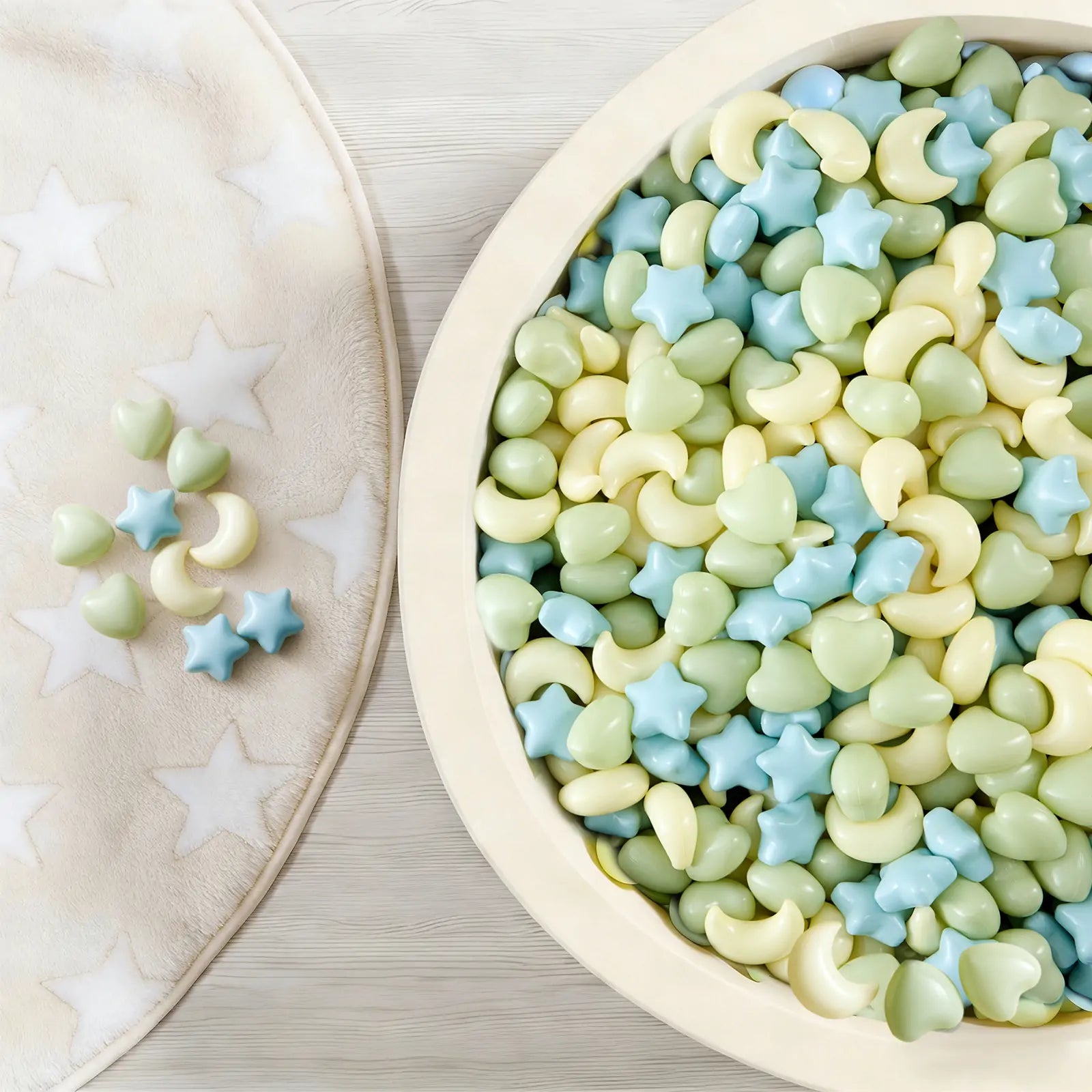 A bowl of pastel star, heart, and moon-shaped candies sits beside a soft white star-patterned blanket—perfect for sensory play or enjoying after fun in the Tiny Land® Green Adventure 3-in-1 Play Tunnel & Fun-Shapes Ocean Ball Set.