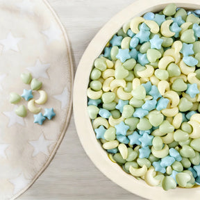 A bowl of pastel star, heart, and moon-shaped candies sits beside a soft white star-patterned blanket—perfect for sensory play or enjoying after fun in the Tiny Land® Green Adventure 3-in-1 Play Tunnel & Fun-Shapes Ocean Ball Set.