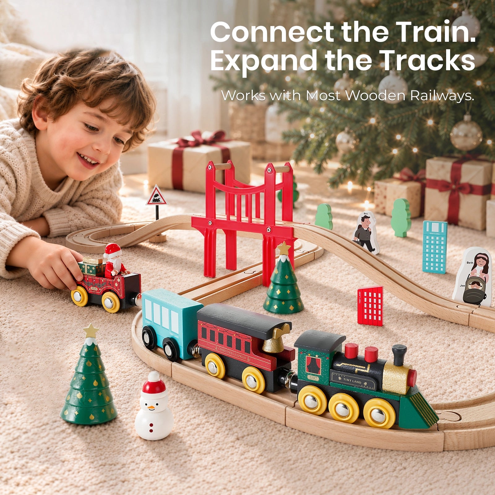 A child plays with a Tiny Land® Christmas Express Wooden Train Accessory Set on a carpeted floor in front of a Christmas tree, with the text Connect the Train. Expand the Tracks. Works with Most Wooden Railways overlaid.