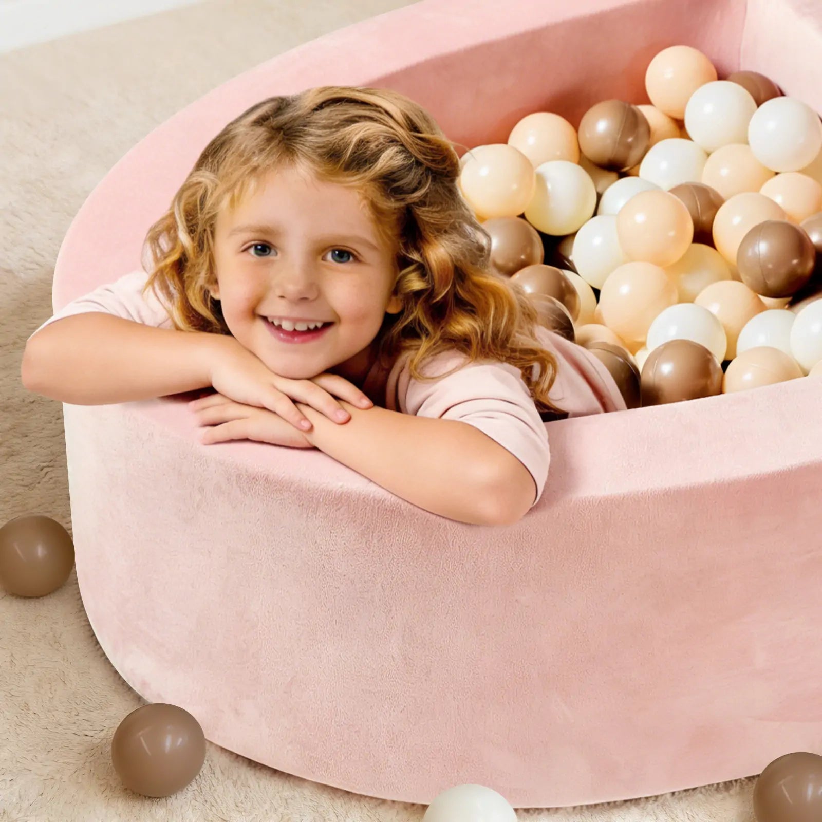 A young girl with wavy blonde hair smiles while leaning on the edge of the Tiny Land® Pink PlayNest Ball Pit by Tiny Land INC, filled with white, beige, and brown balls (Ocean Ball not included). Some balls are scattered on a light carpeted floor.|pink