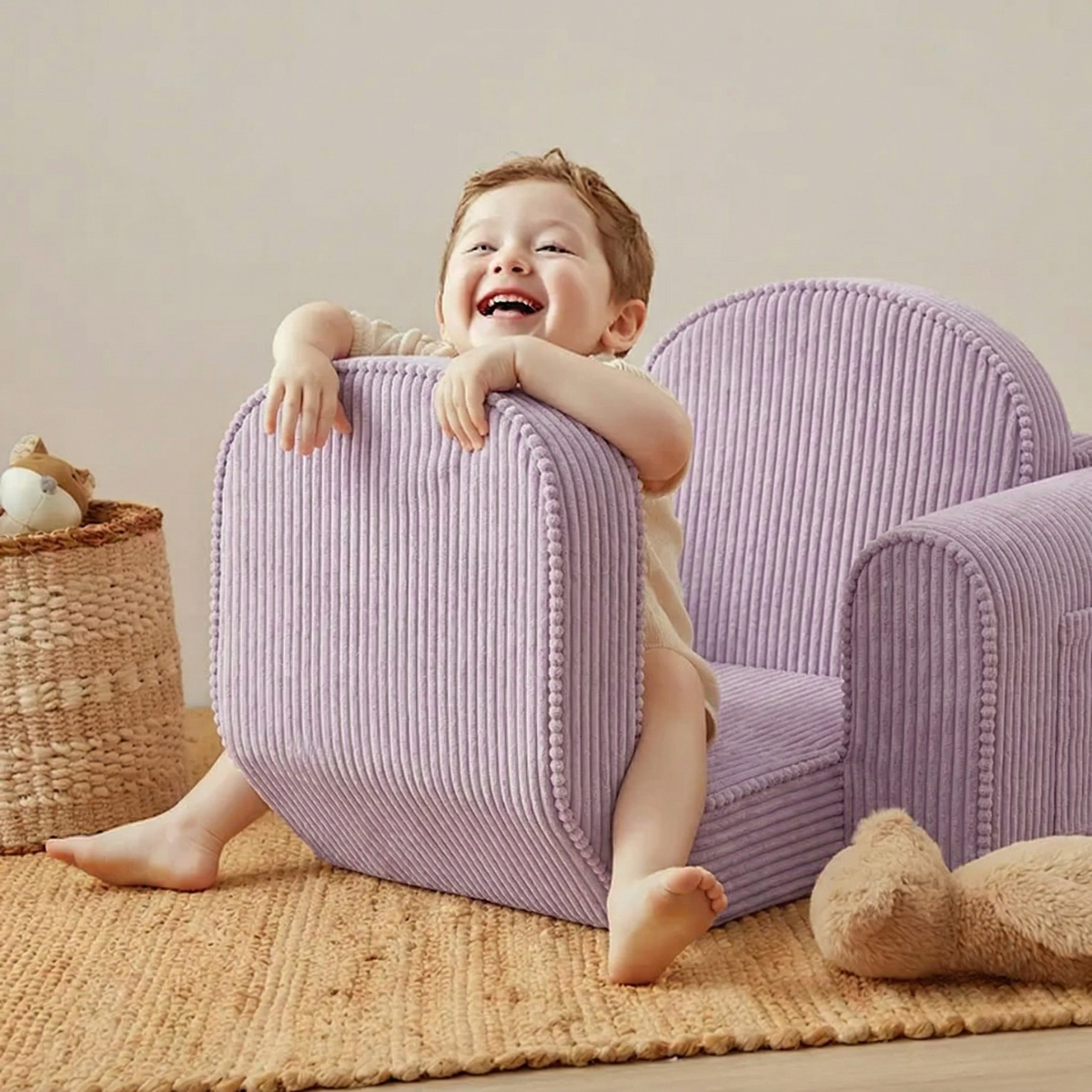A smiling child sits playfully on the Tiny Land® GentleSnug Kid Chair in purple, hugging the backrest. A woven basket with plush toys and a stuffed animal rest on a tan rug nearby, creating a cozy and cheerful scene.