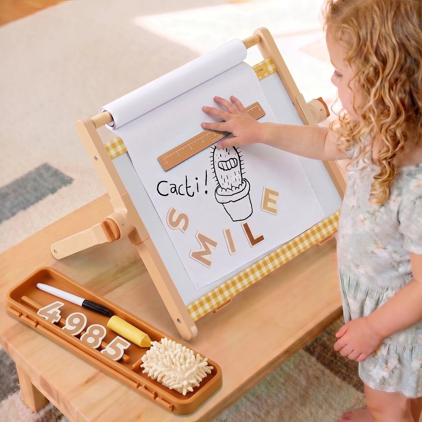 A young child touches a drawing of a cactus and the word SMILE on the Tiny Land® Double-Sided Magnetic Tabletop Easel. Nearby, chalk, an eraser, and number magnets rest in a tray on the wooden table, creating the perfect kids art station by Tiny Land.|Oatmeal