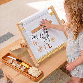 A young child touches a drawing of a cactus and the word SMILE on the Tiny Land® Double-Sided Magnetic Tabletop Easel. Nearby, chalk, an eraser, and number magnets rest in a tray on the wooden table, creating the perfect kids art station by Tiny Land.|Oatmeal