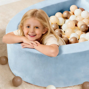 A smiling blonde child leans over the Early Bird Eggs-Tiny Land® PlayNest Ball Pit by Tiny Land INC, filled with 200 white, beige, and brown ocean balls, with some scattered on the beige carpet around the cozy foam nest.|blue