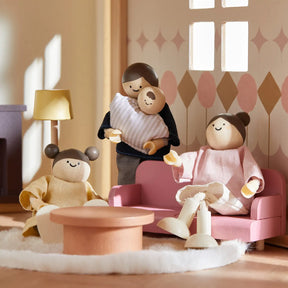 Three Tiny Land® Sweetwood Pegasus Dollhouse wooden dolls—a parent with a baby, another on a pink couch, and a child by the round table—are posed in the sunlit living room with cozy pink furniture.