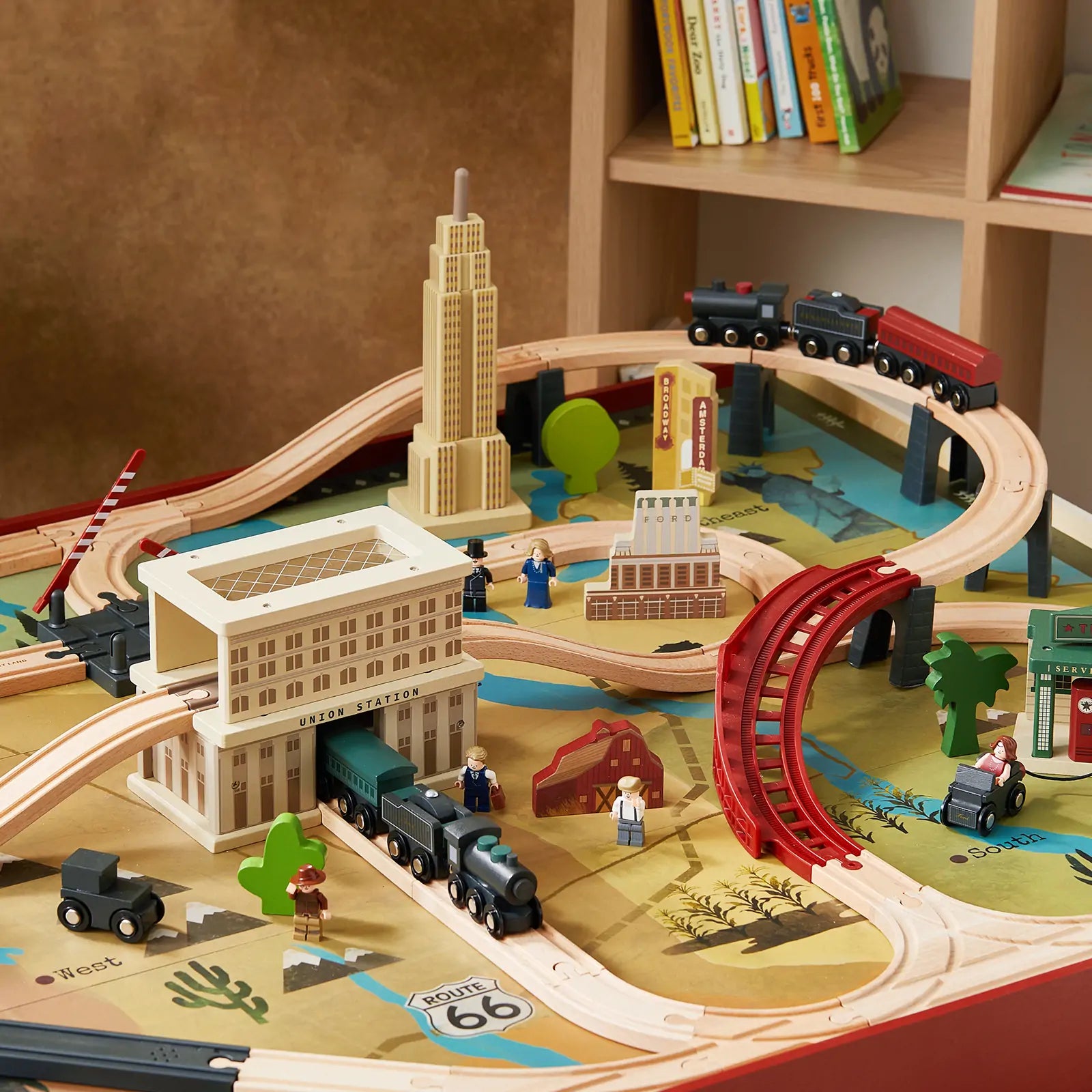 The Tiny Land® 1920 Railway Adventure Train Table by Tiny Land INC features a vintage wooden locomotive set with tracks, bridges, buildings, and accessories, all arranged on a colorful map-themed play table with nearby shelves for books.