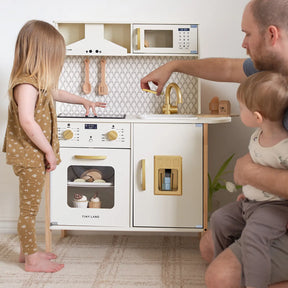A young girl plays at the Tiny Land® PopJoy Play Kitchen, as an adult holding a toddler points at the sink. This Tiny Land set includes miniature utensils, an oven, and a patterned backsplash for imaginative pretend play.