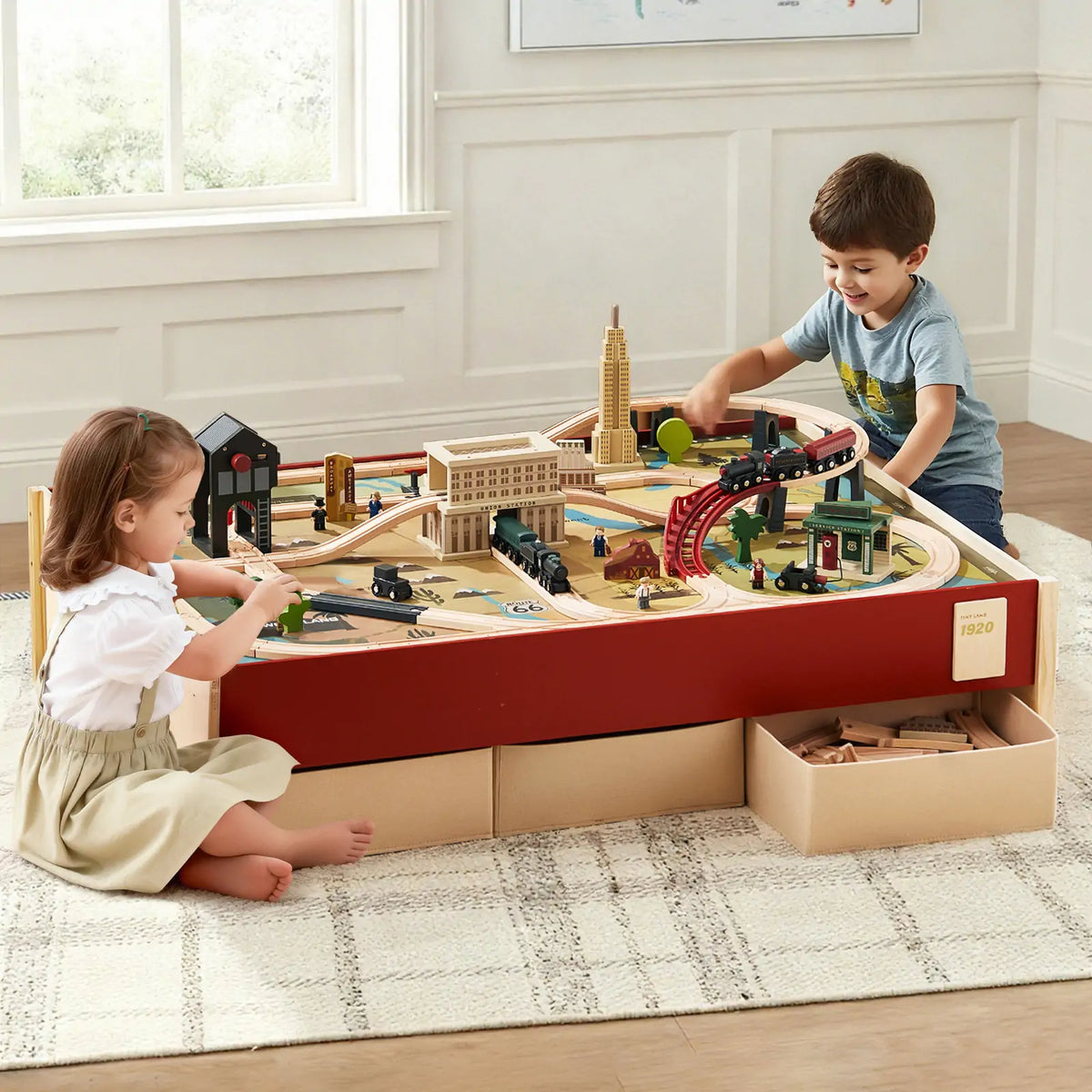 Two children play with the Tiny Land® 1920 Railway Adventure Train Table by Tiny Land. The table has built-in storage drawers and is topped with tracks, buildings, and classic wooden train set pieces in a bright room.