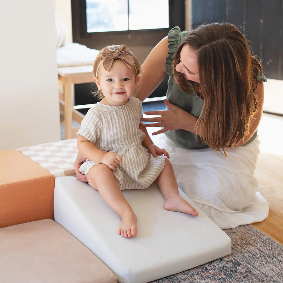 A smiling toddler in a striped dress sits on Tiny Land® PlayNest Climbing Blocks as an adult woman kneels beside her, both smiling in a bright, cozy room designed for gross motor development by Tiny Land.