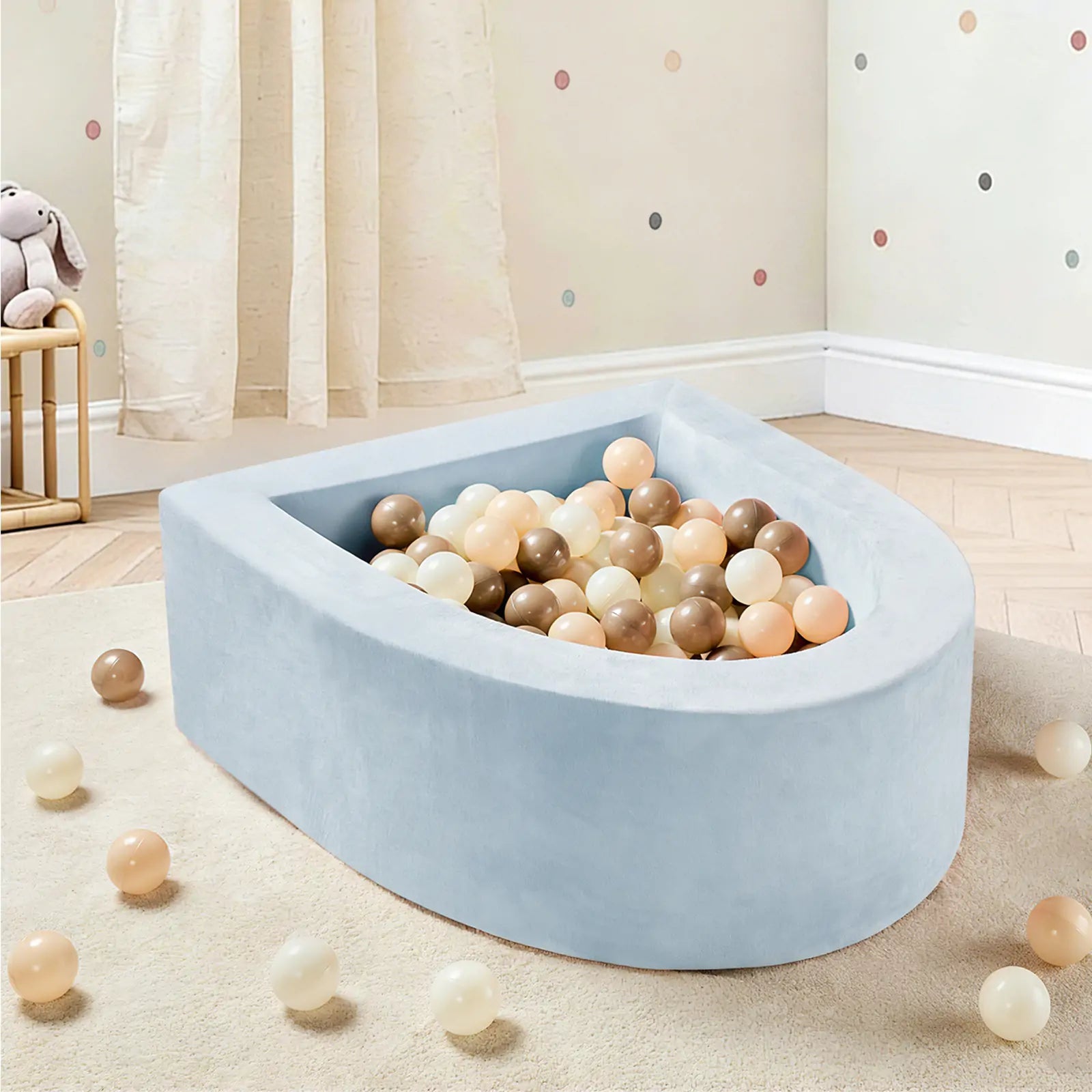 The Tiny Land® PlayNest Ball Pit with 200 Ocean Balls by Tiny Land, featuring a light blue, soft foam design and filled with pastel and neutral-colored ocean balls, sits invitingly in a nursery with wooden floors, polka dot walls, and flowing curtains. A few balls are scattered on the floor, creating a delightful play space for toddlers.|Blue