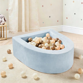 The Tiny Land® PlayNest Ball Pit with 200 Ocean Balls by Tiny Land, featuring a light blue, soft foam design and filled with pastel and neutral-colored ocean balls, sits invitingly in a nursery with wooden floors, polka dot walls, and flowing curtains. A few balls are scattered on the floor, creating a delightful play space for toddlers.|Blue