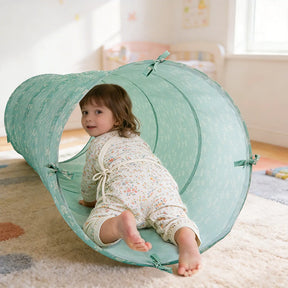 A young child in floral pajamas crawls through the Tiny Land® Adventure Play Tunnel - Green by Tiny Land in a cozy, sunlit room with a rug and toys in the background.|green