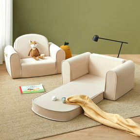 A cozy kids’ room features the Tiny Land® GentleSnug Kid Chair - Beige White, a stuffed animal, a foldable kids sofa bed with a yellow blanket, wooden blocks, a book on the rug, and a green basket against calming green walls.