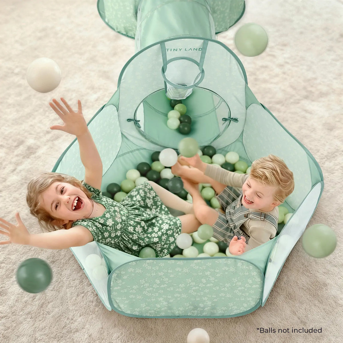 Two young children play and laugh in the mint green Tiny Land® Compact Adventure 3-in-1 Play Tunnel, tossing white and green balls. The ball pit features soft pastel colors and a floral pattern. Text reads: "*Balls not included.
