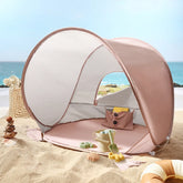 The Tiny Land® One-Stop Beach Toy & Tent Set – Blush Pink features a small UV-protective tent and sand play set, perfect for the beach. It sits on sandy ground by the ocean, with a basket and decorative shells nearby under the blue sky.