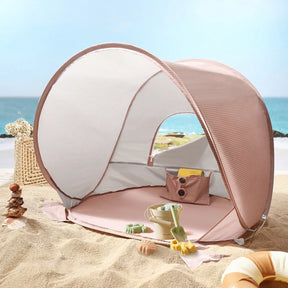 The Tiny Land® One-Stop Beach Toy & Tent Set – Blush Pink features a small UV-protective tent and sand play set, perfect for the beach. It sits on sandy ground by the ocean, with a basket and decorative shells nearby under the blue sky.