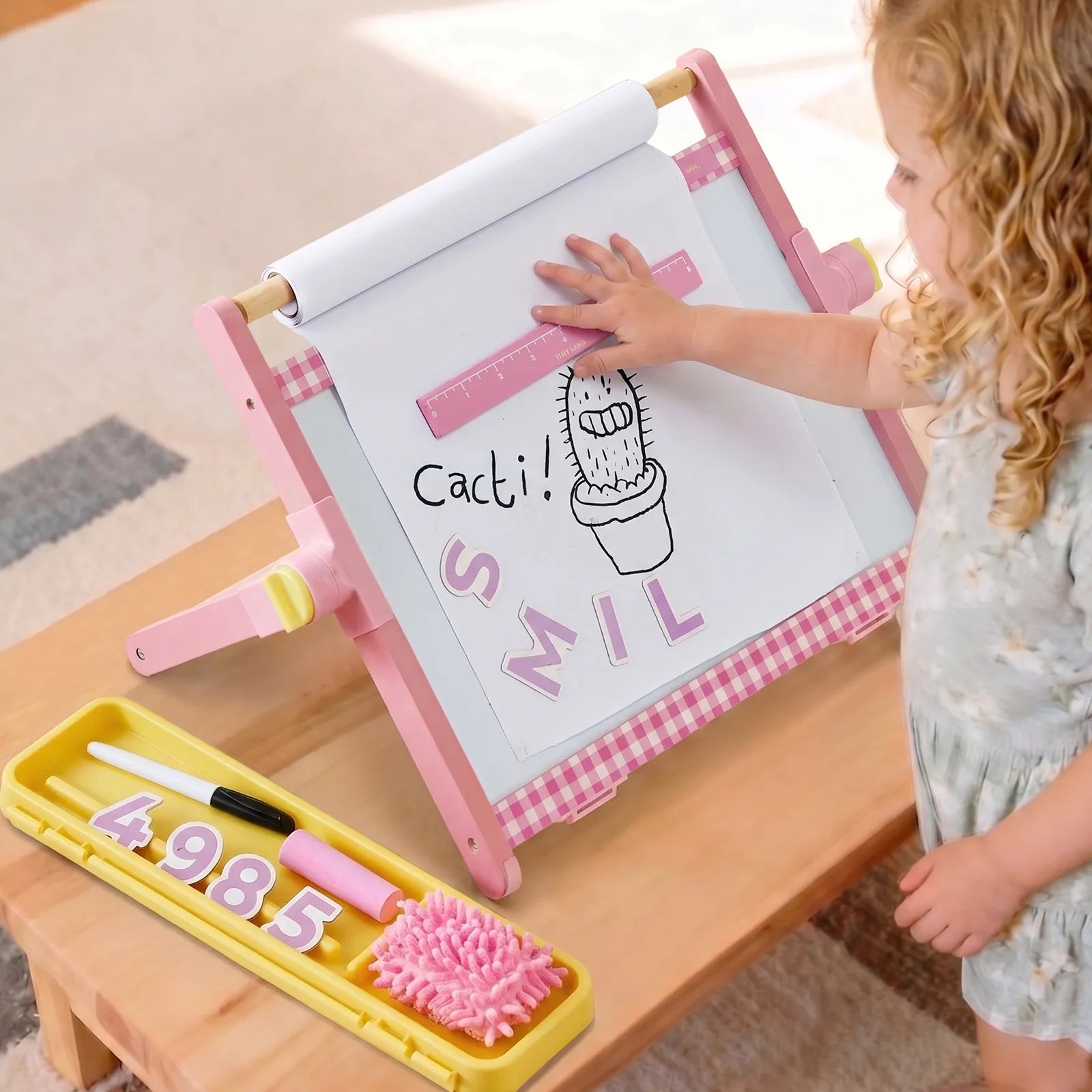 A young child uses the Tiny Land® Double-Sided Magnetic Tabletop Easel by Tiny Land as their kids’ art station, holding a ruler over a cactus drawing labeled “Cacti! SMILE.” Art supplies, numbers, and a duster rest on the nearby yellow tray atop the wooden table, while the magnetic whiteboard side provides extra creative space.|Pink