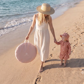 A woman in a white dress and sunhat walks barefoot on the beach, holding hands with a toddler in pink. Their Tiny Land® 4-in-1 Baby Beach Tent & Sand Toy Set provides shade for play as waves gently touch the shore nearby.|Pink
