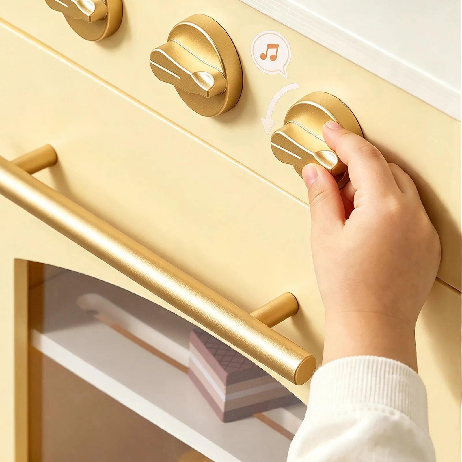 A child’s hand turns a gold-colored knob on the Tiny Land® Wooden Play Kitchen with Storage for Creative Toddlers by Tiny Land. A music note symbol indicates that the knob produces sound. The oven door features a gold handle and opens to reveal a shelf with pretend cookware and toy food, showcasing the play kitchen’s multifunctional storage design.|Pale Yellow
