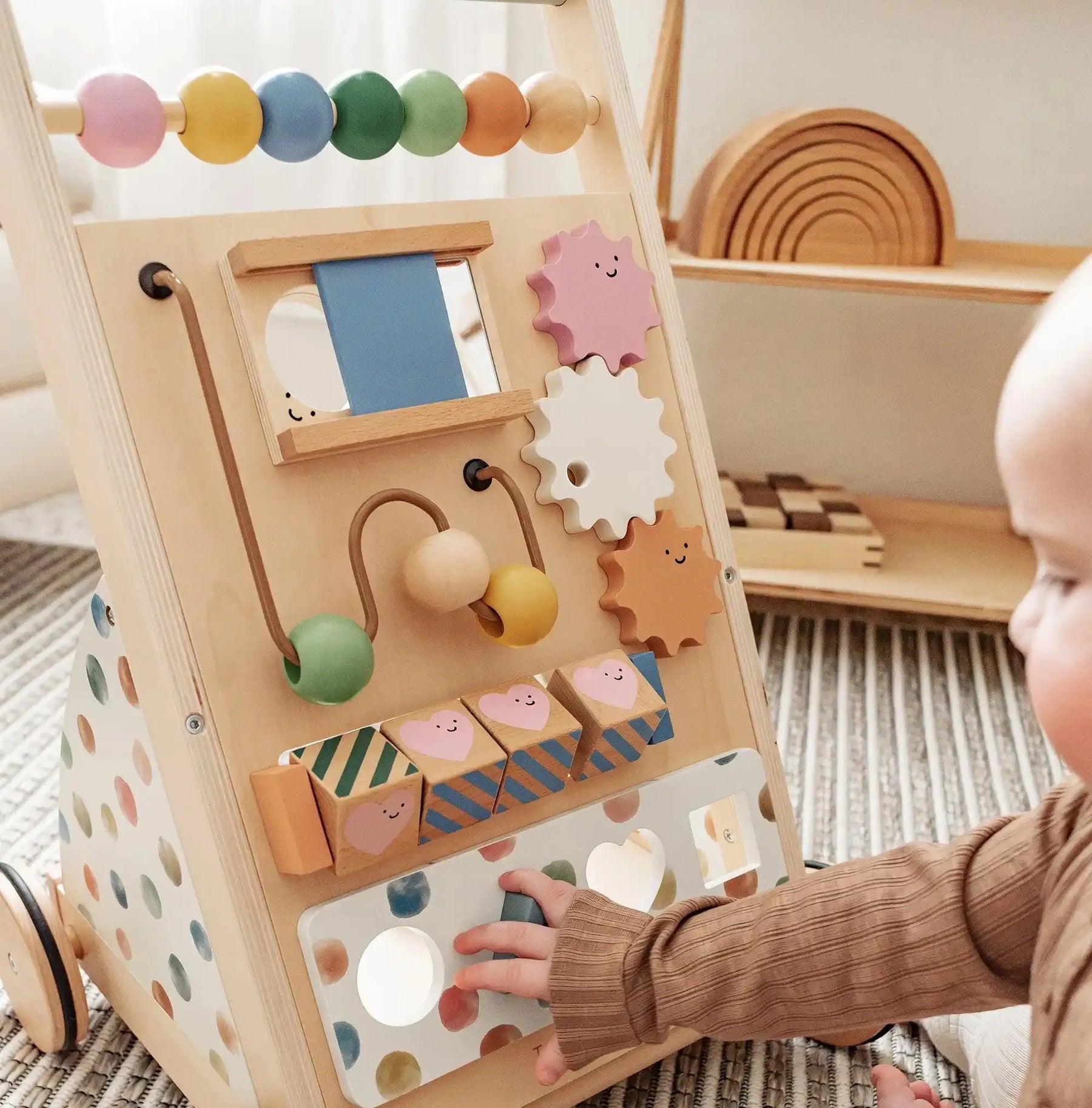 A baby plays with the Tiny Land® Versatile Natural Wooden Baby Walker by Tiny Land, featuring beads, gears, a mirror, and pastel-colored blocks, on a patterned rug in a bright, cozy room.