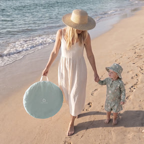 A woman in a sunhat and white dress walks with a toddler along the beach, carrying the Tiny Land® 4-in-1 Baby Beach Tent & Sand Toy Set—the perfect accessory for sand and water fun as ocean waves roll in behind them.|Green