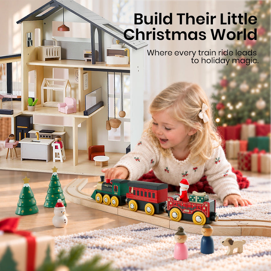 A young child plays with a Tiny Land® Christmas Express Wooden Train Accessory Set on a track in front of a dollhouse, with two small Christmas trees and figures of a snowman, people, and a dog nearby.