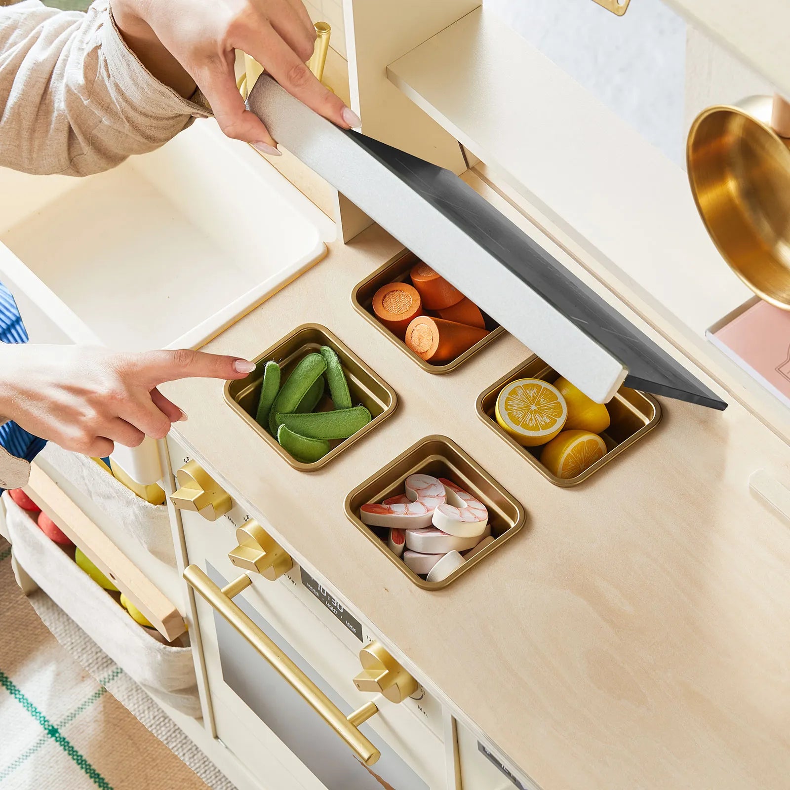 A child lifts the lid of the Tiny Land® BistroSwitch Play Kitchen by Tiny Land, revealing three compartments with toy veggies and fruits—snap peas, carrot slices, lemon halves, and pink-and-white swirled pieces for pretend play.|White