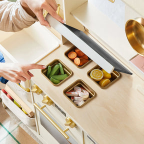 A child lifts the lid of the Tiny Land® BistroSwitch Play Kitchen by Tiny Land, revealing three compartments with toy veggies and fruits—snap peas, carrot slices, lemon halves, and pink-and-white swirled pieces for pretend play.|White