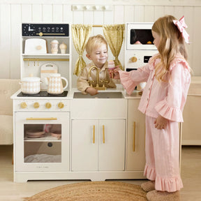 Two young children play with the Tiny Land® Iconic Wooden Play Kitchen – Cream Pretend Cooking Set for Kids. The boy stands behind the sink, while the girl, dressed in pink ruffled pajamas, stands in front holding a small item. This Tiny Land play kitchen features elegant gold accents and patterned utensils, inspiring hours of imaginative pretend cooking and creative play.