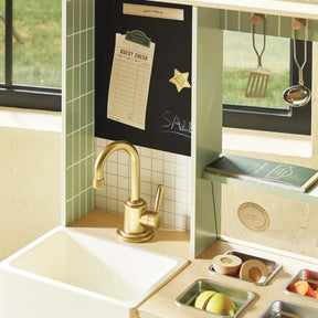 The Tiny Land® BistroSwitch Play Kitchen features a gold faucet, white sink, chalkboard with "SALE," toy utensils, and pretend food items. Sunlight streams in, making this Tiny Land set ideal for imaginative pretend play.|Green