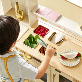 A young child in an apron uses tongs to pick up toy spinach, bread, red cubes, and orange pieces from metal trays while playing at the Tiny Land® BistroSwitch Play Kitchen by Tiny Land.|White