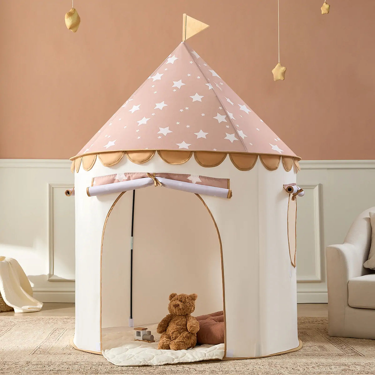 The Tiny Land® Cheerful Pennant Castle Tent features a pink star-patterned roof and scalloped trim. Inside, a brown teddy bear rests on a cushioned mat, with beige walls and hanging star décor creating a cozy indoor space.