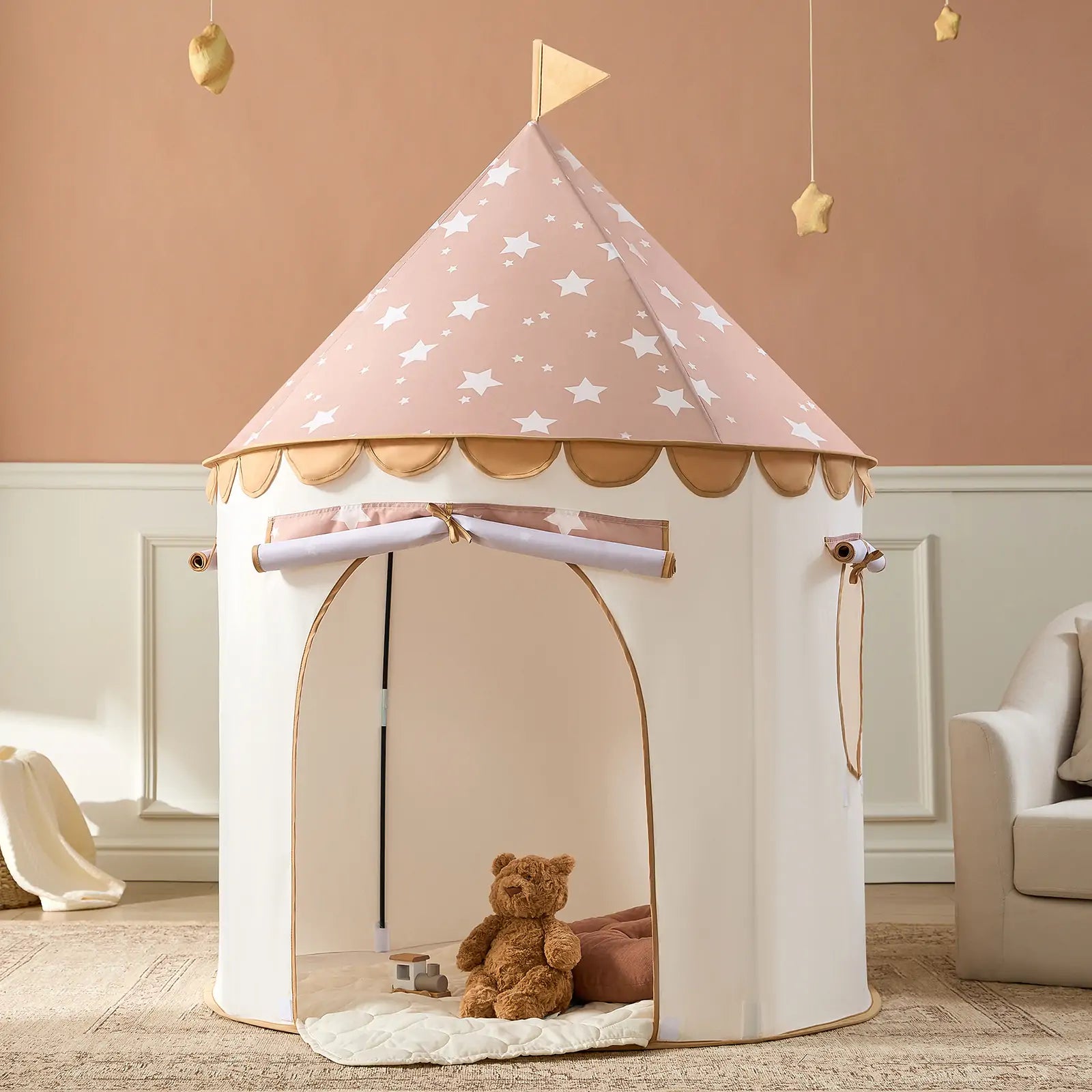 The Tiny Land® Cheerful Pennant Castle Tent features a pink star-patterned roof and scalloped trim. Inside, a brown teddy bear rests on a cushioned mat, with beige walls and hanging star décor creating a cozy indoor space.