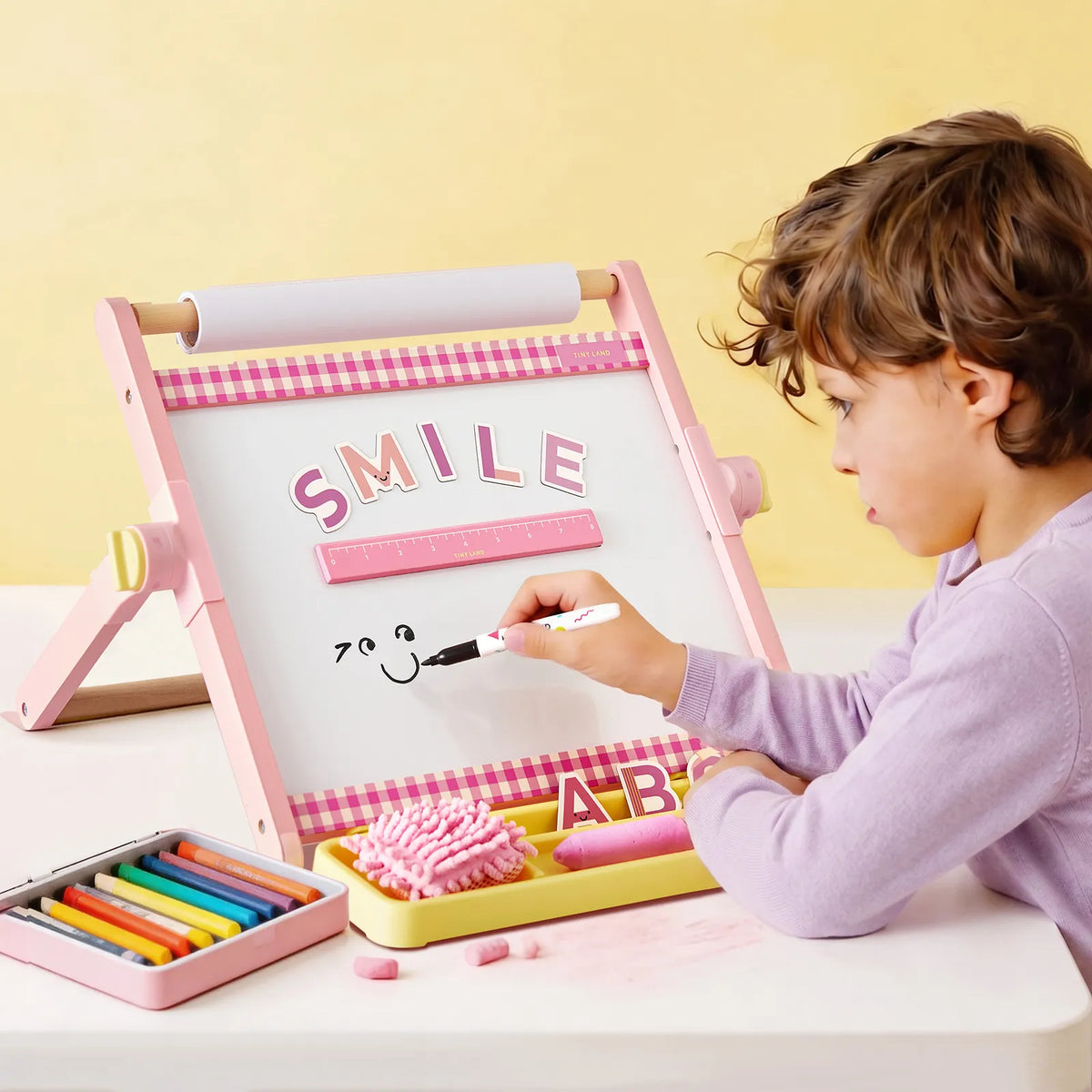 A child in a purple shirt draws a smiley face on the pink and white Tiny Land® Double-Sided Magnetic Tabletop Easel, which features the word "SMILE." Colorful markers and chalk from Tiny Land are nearby, making this branded art station perfect for creative fun.|Pink