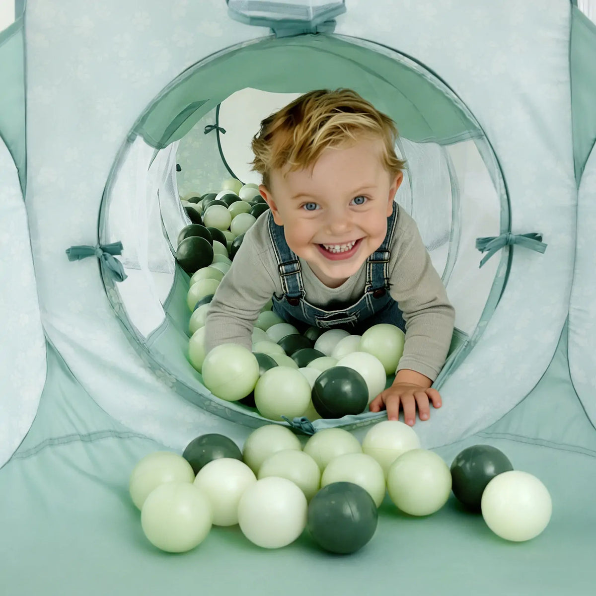 A young child with blonde hair and overalls smiles while crawling through the Tiny Land® Adventure 3-in-1 Play Tunnel & Ocean Ball Set - Green, filled with light and dark green plastic balls by Tiny Land.