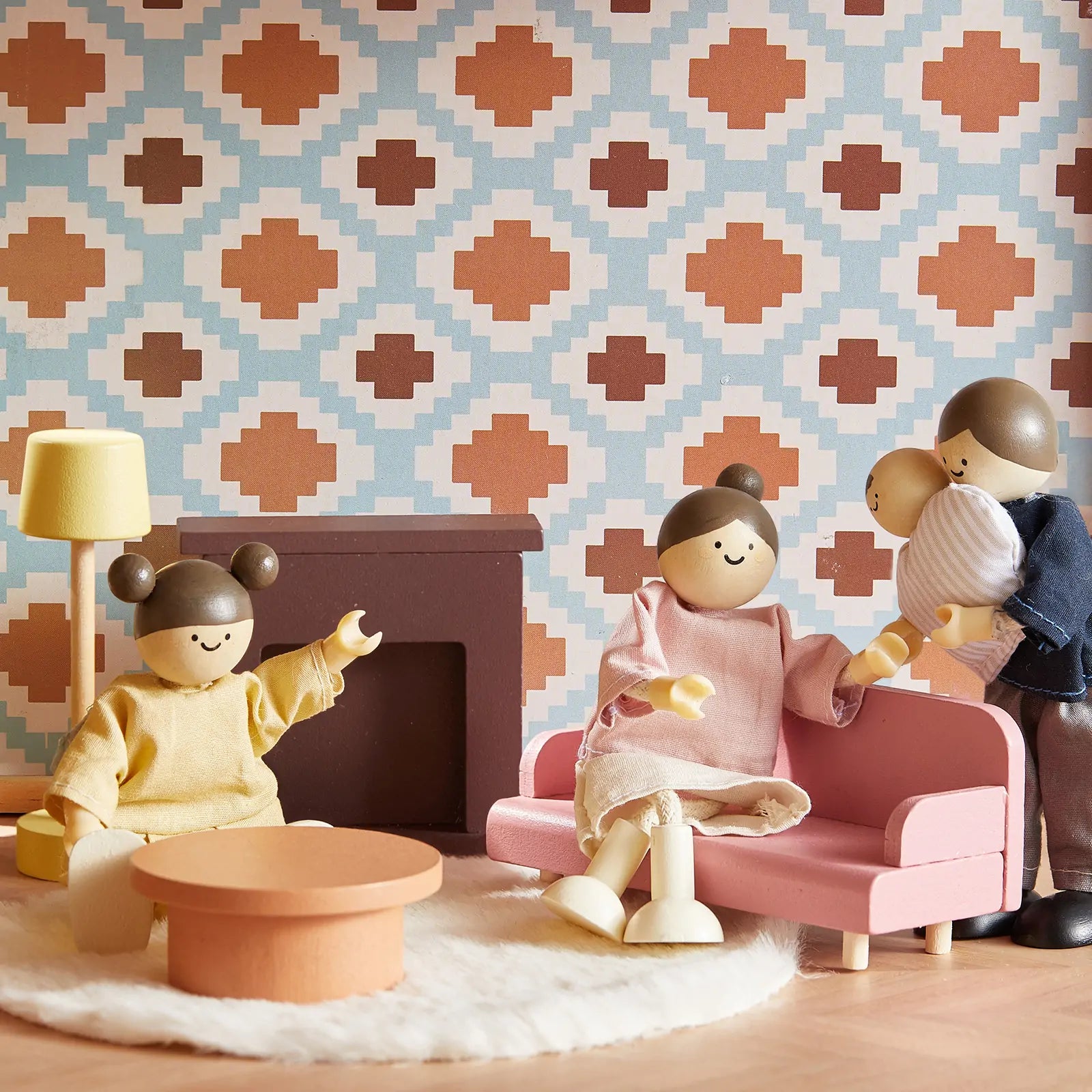 Three Tiny Land® Sweetwood Pegasus Dollhouse wooden dolls—a seated adult, a baby by the fireplace, and a waving child—enjoy the living room with geometric wallpaper and pink couch from the Tiny Land family doll set.
