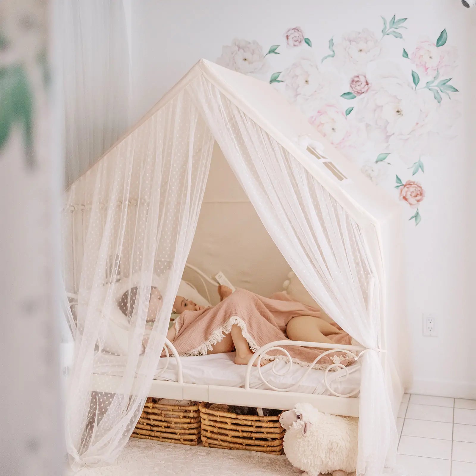 A child rests under a pink blanket in the Tiny Land® Large Space Play House with Star Lights by Tiny Land, featuring sheer curtains. The room includes floral wall art, storage baskets, and a plush sheep toy on the white tiled floor.