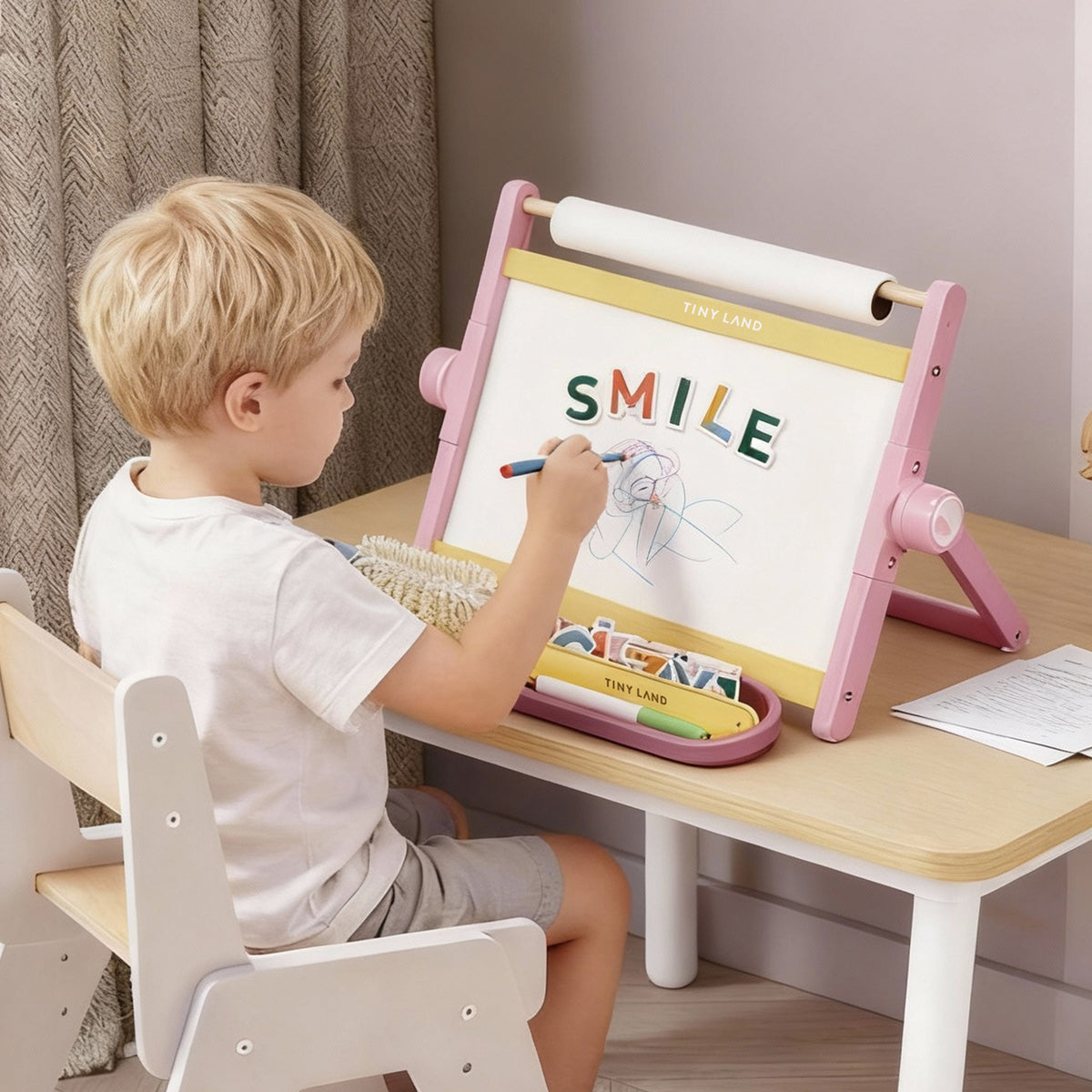 A young boy draws on the Tiny Land® Double-Sided Magnetic Tabletop Easel - Pink, with "SMILE" in colorful letters above his art and supplies neatly arranged in a tray below, from the brand Tiny Land.