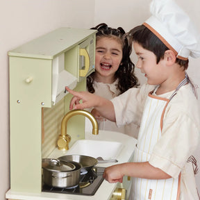 Two young children play with the Tiny Land® PopJoy Play Kitchen - Green. The boy, dressed as a chef, presses a button while the girl smiles behind him. Pots and utensils from Tiny Land are on the stovetop.