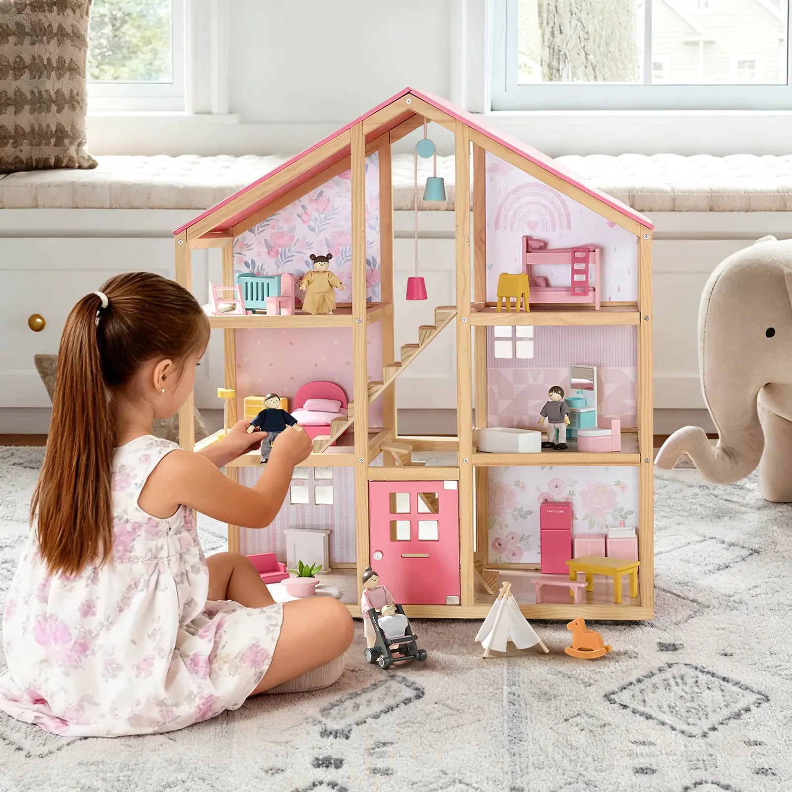 A young girl in a floral dress enjoys imaginative play with the Tiny Land® Sweetwood Love Dollhouse & Wooden Dollhouse Dolls by Tiny Land INC, arranging the set near her favorite stuffed elephant on a patterned rug.