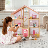 A young girl plays with the Tiny Land® Sweetwood Love Dollhouse & Wooden Dollhouse Dolls, arranging furniture on a patterned rug beside a stuffed elephant in a bright, cozy room.